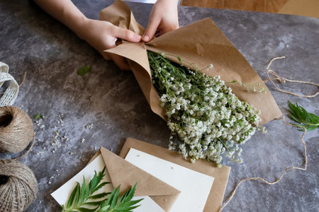 Florist at work: a pretty woman prepares a summer bouquet of white gypsophila on a gray desk. Kraft...
