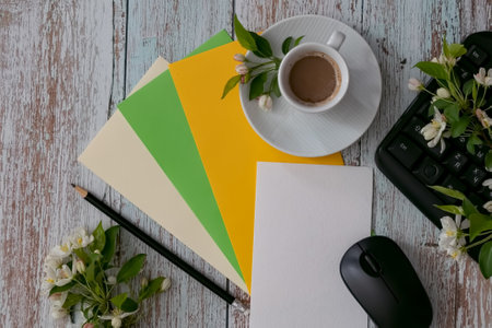 A cozy workspace features a black notebook with a pencil, a cup of coffee on a white saucer, and delicate flowers, alongside a computer keyboard and mouse on a wooden table.の写真素材