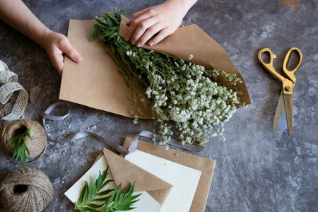 Hands arrange delicate white flowers with twine and decorative ribbons on a rustic surface,...