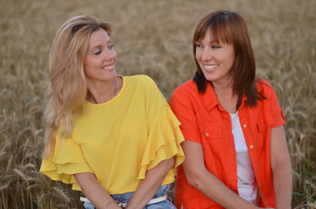Two smiling women sit closely together in a wheat field at sunset, sharing laughter and enjoying the warm evening atmosphere in bright, colorful clothing.の写真素材