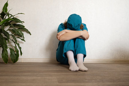 A healthcare worker wearing scrubs and a cap sits with their knees hugged to their chest, showing signs of emotional exhaustion. The serene environment contrasts with their apparent distress.の写真素材
