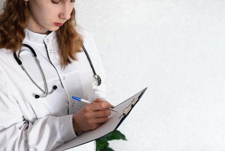 A young medical professional in a white coat writes on a clipboard, focused on patient information....