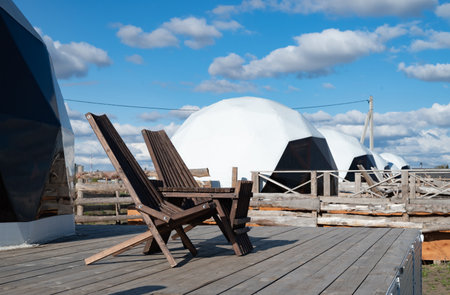 A wooden reclining chair sits on a deck, providing a perfect view of modern dome structures. The scene is set against a clear blue sky with fluffy clouds, creating a tranquil atmosphere.の写真素材