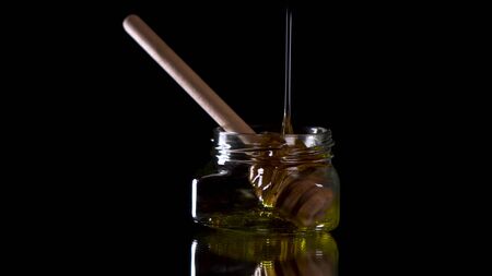 Pouring honey in transparent jar with honey spoon inside on black background.の写真素材