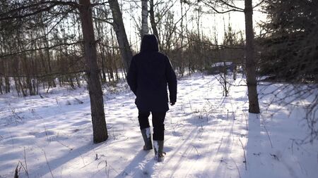 lonely man in blue jacket and high boots walking on snowy field, sun is shining into camera,...