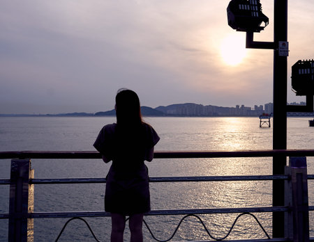 A silhouette of an asian woman standing beside a pole at the edge of a park near the ocean at...