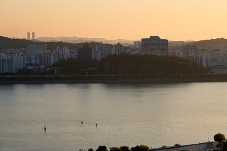 A peaceful wide-angle view of sailboats drifting on the Han River at Haneul Park in Seoul, South Korea, during a hazy golden sunset with the city skyline and mountains in the distaの写真素材