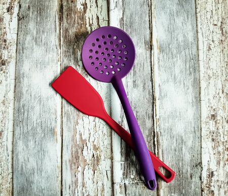 Kitchen utensils. Red plastic kitchen spatula and violet plastic slotted spoon on background with aged wooden boards.の写真素材