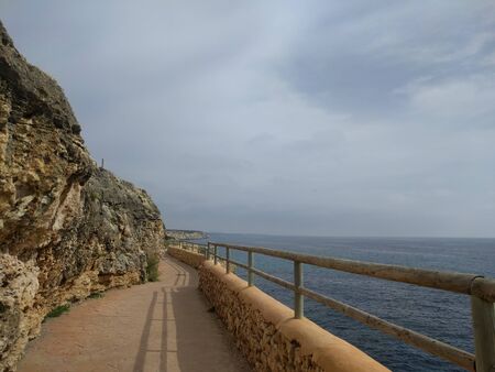 Boardwalk along the sea along a high rocky shore with a wooden fence. Romantic summer walk in the rays of the setting sun. Romantic travel concept. Spain, island of Mallorca.の写真素材