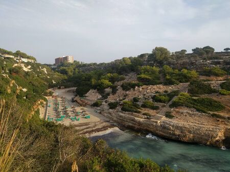 View from the high rocky shore to the sandy beach in the bay of Cala Antena on the island of Mallorca, Spain. A deserted beach with parasols and beach chair in the rays of the rising sun.の写真素材