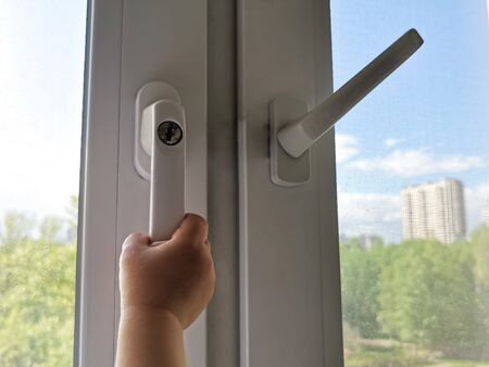 A children's hand is trying to open a window with a lock on the handle in an apartment on a high floor. A security lock for windows ensures the safety of your children and prevents accidents.の写真素材