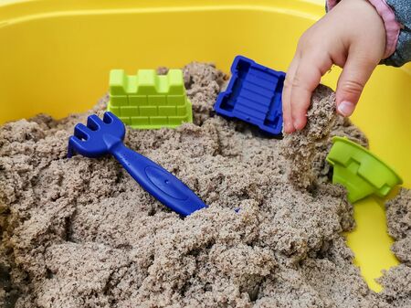 A child plays with kinetic sand: closeup of a child's hand and sand. Sand in a yellow container with plastic blue toy rake and other toys.の写真素材