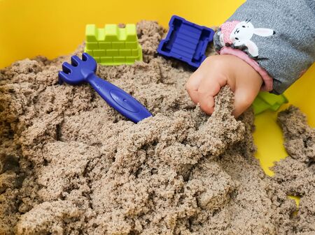 A child plays with kinetic sand: closeup of a child's hand and sand. Sand in a yellow container with plastic blue toy rake and other toys.の写真素材