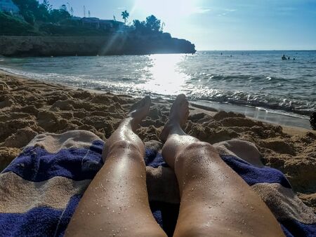 Woman lies on a towel on a sandy beach opposite the bright sun. The legs are located towards the sea, on the feet are a drop of water. In the background is a rocky shore and blue sky. Summer vacationの写真素材