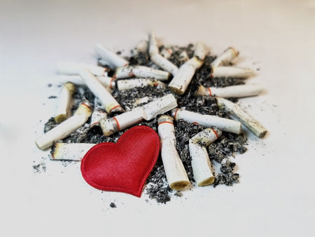 Closeup red heart lies on a pile of cigarette butts and ashes on a white background. Symbol of the...