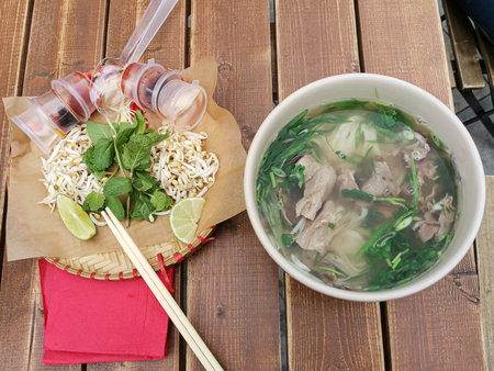 Lunch at a Vietnamese street cafe. A deep plate with beef broth, herbs and meat and a plate with...