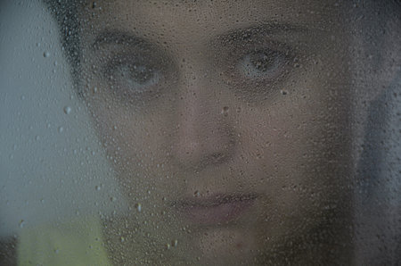 Portrait of a young brunette woman behind glass with rain drops. The concept of depression, blurring...