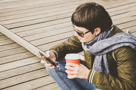 Young brunette woman with short hair in jeans and jacket sits on wooden pier with cup of coffee in...