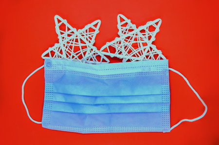 Red Christmas background with two white Bethlehem straw stars under a blue medical mask in the...