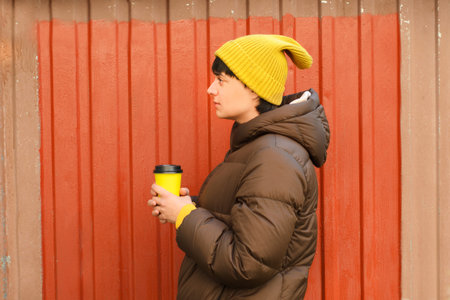 Young woman in profile in yellow knit hat and autumn clothes with cup of coffee in hands against...