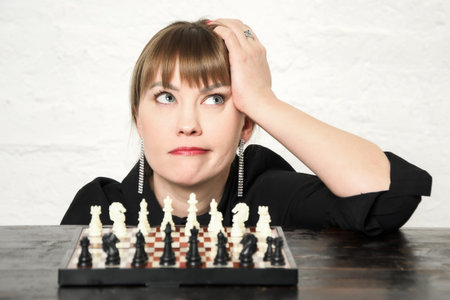 Beautiful young woman is looking pensively upwards while sitting in front of chessboard with spaced...