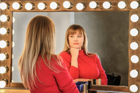 Young woman with blond hair in red on dressing table looks thoughtfully at her reflection in the...