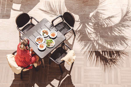 Woman in red dress sits at a table in sun under shade of palm trees. There are several plates with a variety of food on the table. All inclusive rest. Top viewの写真素材