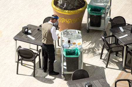 Evrenseki, Turkey - 03.17.2021 Waiter in medical mask and plastic visor clears dirty dishes from...