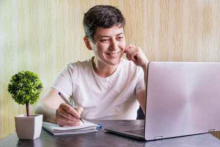 Brunette woman with short hair sits at a table in front of a laptop with a notebook and a pen. Woman...