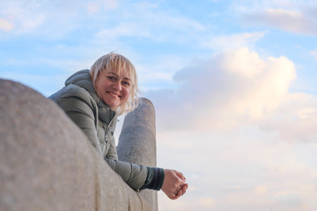 Blonde middle-aged woman stands at stone railing against a background of blue sky. A woman with hair...