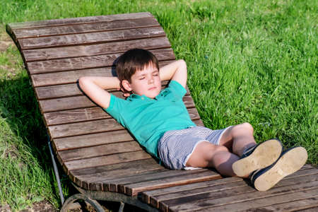 Brunet boy of 8 years in T-shirt and shorts lies on wooden deck chair in the sun against the backdrop of a green lawn. The boy in relaxed, lazy position, closed his eyes.の写真素材