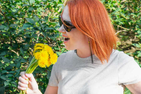Young woman with red hair in sunglasses fooling around portrays singing with a microphone holding a bouquet of yellow dandelions in her hand. Walks in the park, spring mood, fun.の写真素材