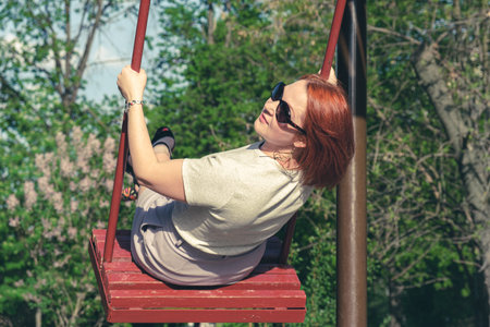 Young woman with red hair in sunglasses smiles swinging on a swing in a city park. The woman on the swing turned around while moving. Childrens entertainment for adultsの写真素材
