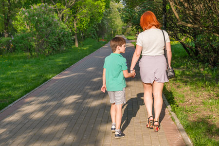 Young woman with red hair in shorts and her son are walking along path of city park. Photographed...