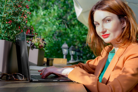 Red-haired young woman in a elegant blazer sits on the open veranda of a cafe with a laptop. Working...