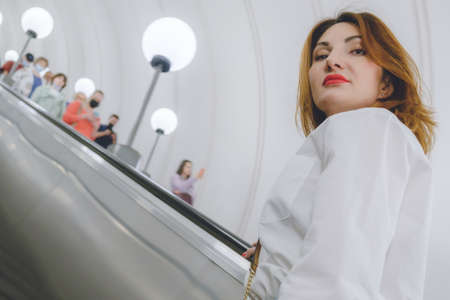 Attractive young woman in a white shirt and with bright red lips rides up the escalator in the subway. Woman looks up haughtily.の写真素材