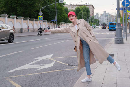 Attractive red-haired young woman in a red baseball cap and beige raincoat stands on the side of a city road and get a taxi. Walking around the city.の写真素材