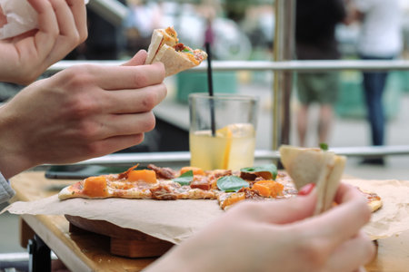 Close-up of hands and pizza with basil leaves in an outdoor street cafe. People eat pizza with their...