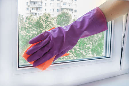 Woman's hand in a lilac rubber glove wipes a glass unit window in a room with a rag. The concept of cleaning company services, window cleaning, housework.の写真素材