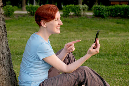 Young red-haired woman with a short haircut sits on the grass near a large tree and talk via video communication on a smartphone. The woman laughs and points her index finger to the screenの写真素材