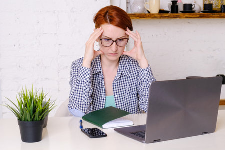 Young red-haired woman with glasses sits at table in front of a laptop with her fingers to her...