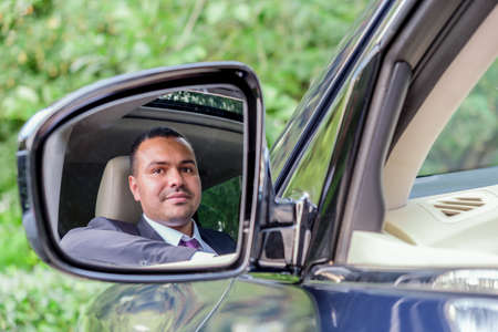 Young man of Middle Eastern appearance in a business suit sitting behind the wheel of a car looks at his reflection in the side rearview mirror. Businessman driving his car. Portrait of a driverの写真素材