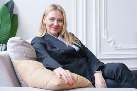 Attractive blonde woman in a business gray suit is sitting on the couch. The woman smiles and looks into the camera. Beautiful businesswoman waiting for a business meeting.の写真素材