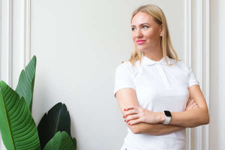 Attractive blonde woman in a white polo shirt stands with her arms folded on her chest against the background of a white wall. Successful female manager looks to the side.の写真素材