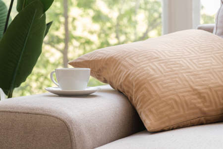 White coffee cup and saucer sits on the arm of the living room sofa on the background of a window with green trees. Concept of a morning cup of coffee, a good start to the day, a cozy home.の写真素材