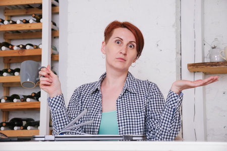 Young woman with short hair, holding a mug in her hand, throws up her hands in surprise. Woman in at...