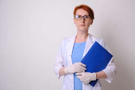 Young red-haired female medic in white coat with a folder for notes in hand. Portrait of a female doctor on gray monochrome background. Doctor looks into cameraの写真素材