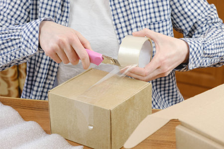 Warehouse employee wraps the order of online store with scotch tape in cardboard box. Woman holds...