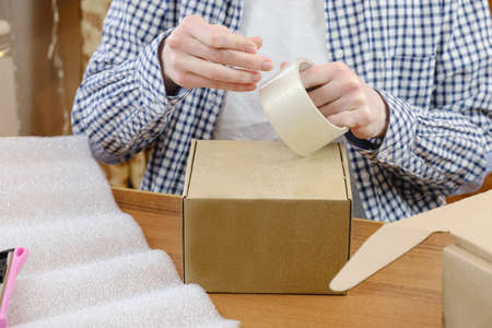 Warehouse employee wraps the order of online store with tape in cardboard box. Small business, order processing and packingの写真素材