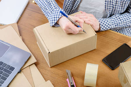 Woman at the desk marks the e-commerce order packed in a cardboard box with a pen. Warehouse employee processes and packs order for delivery, warehouse goods logistics.の写真素材
